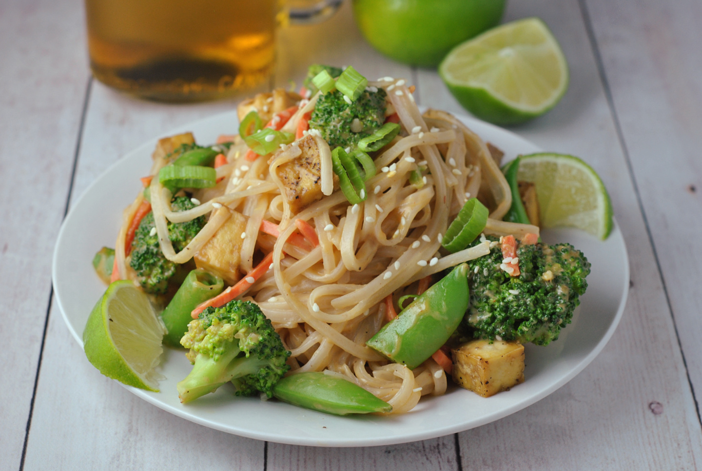 Close-up image of peanut sauce rice noodles with bright green broccoli, snap peas, and wedges of fresh limes. 