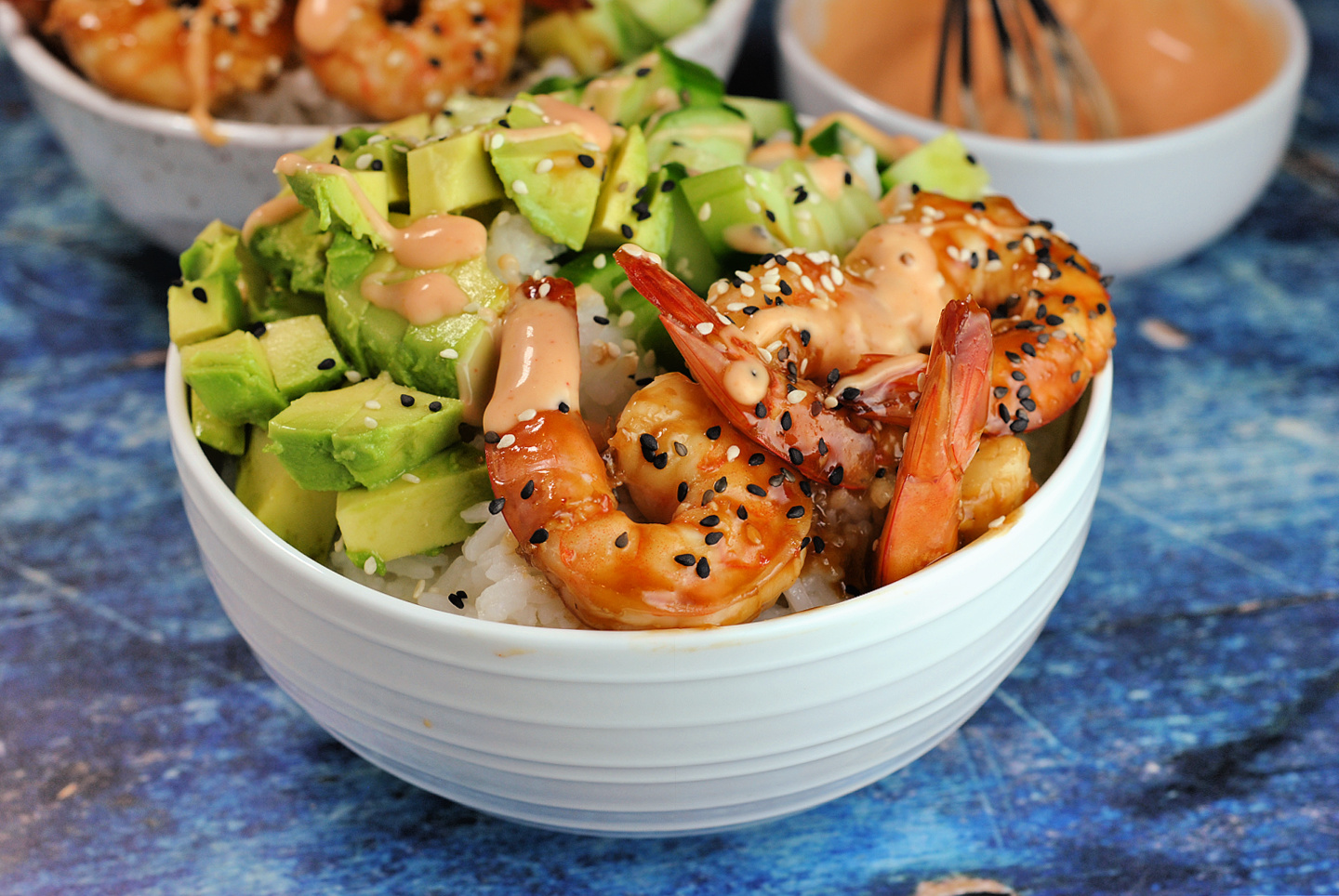 Shrimp rice bowl topped with avocado, cucumber, and sriracha mayo sitting on a blue table. 