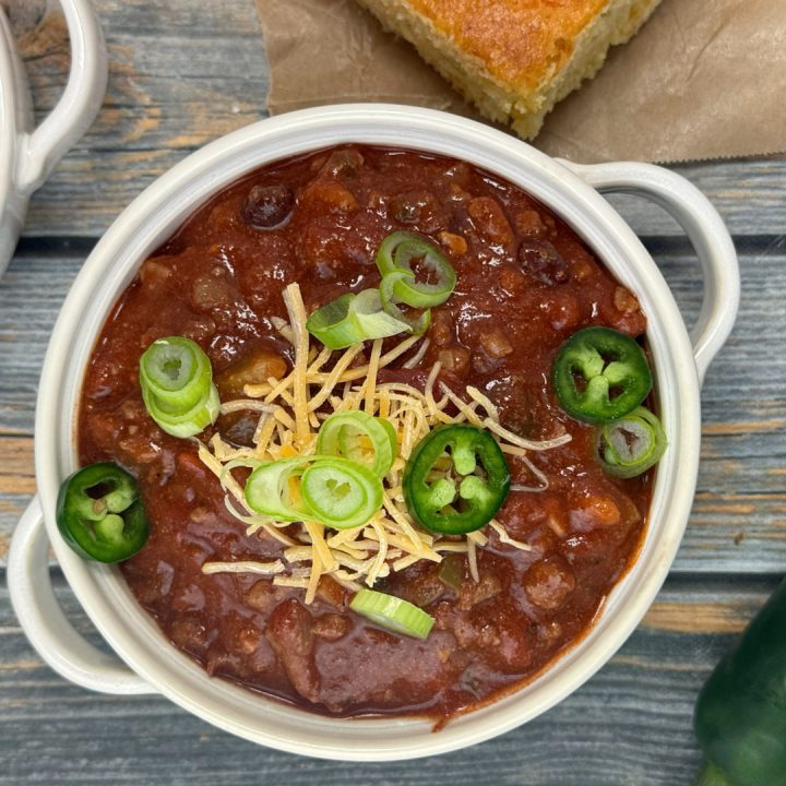 A blue wooden table with a bowl of Beef and Bean Chili topped with shredded cheddar cheese, green onions, and jalapeno slices with a piece of cornbread.