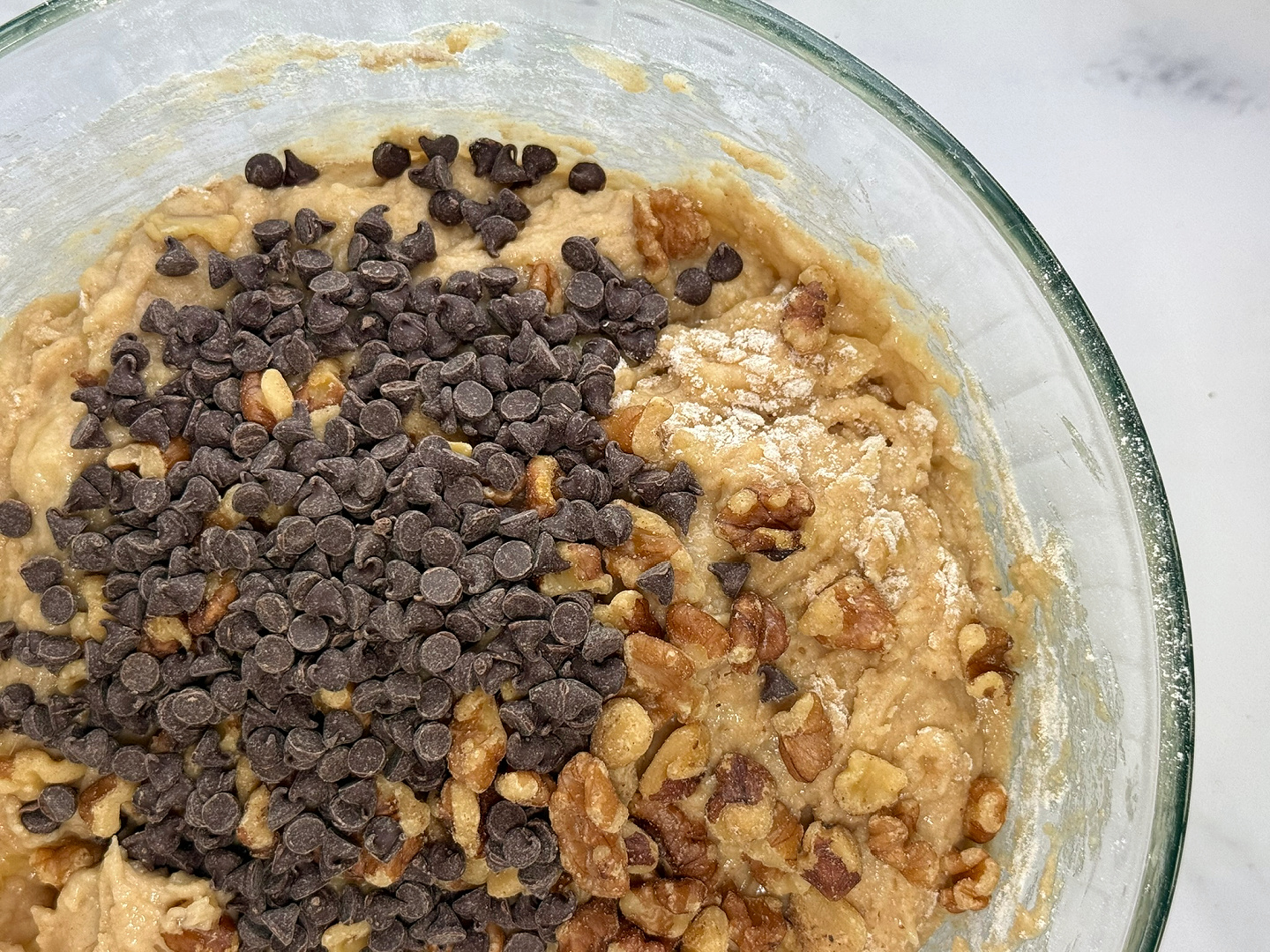 Top-down view of banana bread batter in a clear glass mixing bowl, with a generous pile of chocolate chips and chopped walnuts added on top. The thick batter is partially mixed, with visible streaks of flour along the sides of the bowl.