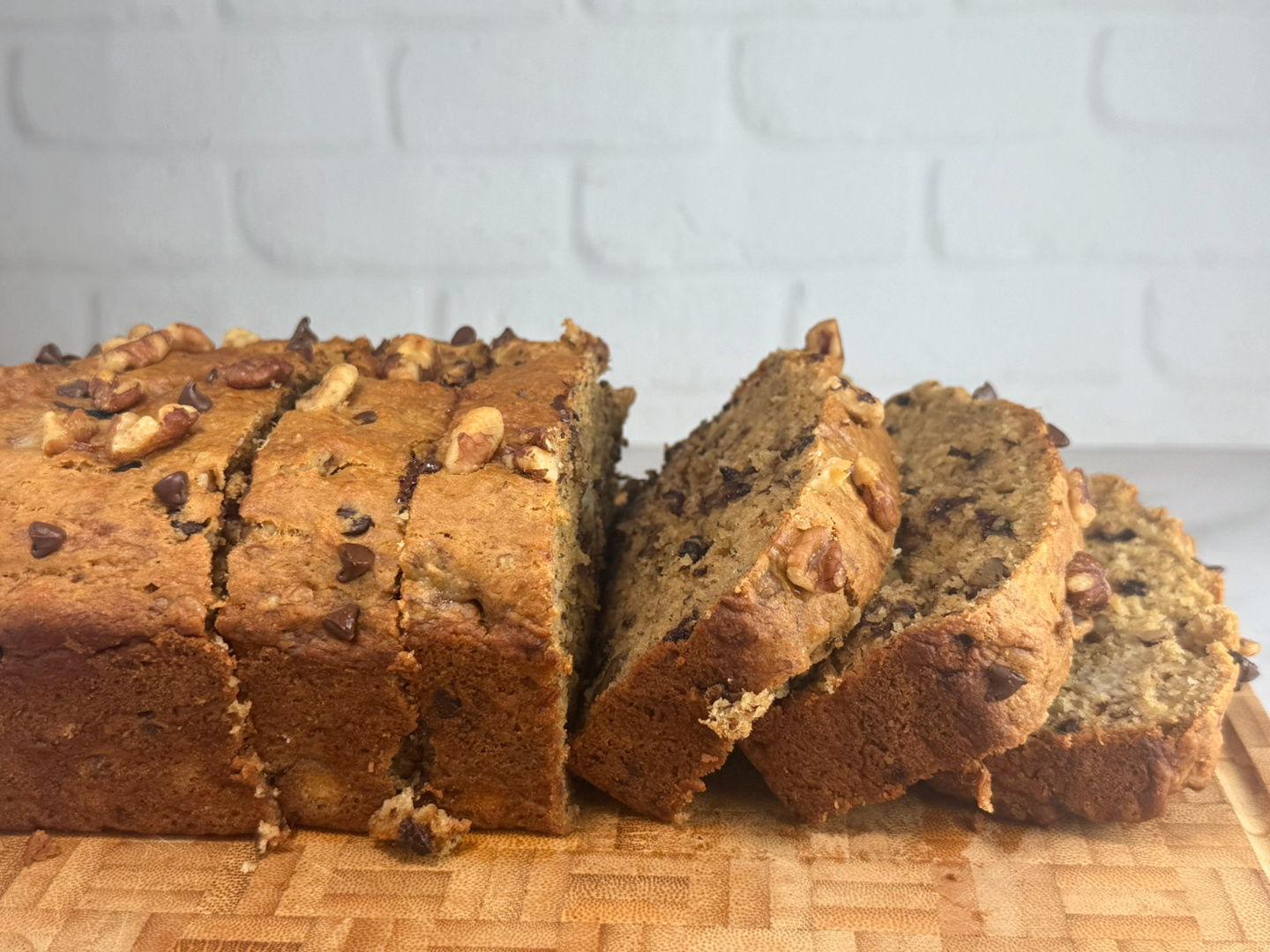 Sliced whole wheat chocolate chip banana nut bread arranged on a wooden cutting board. The loaf has a golden-brown crust with visible walnuts and chocolate chips on top, and the interior appears moist and studded with nuts and melted chocolate.