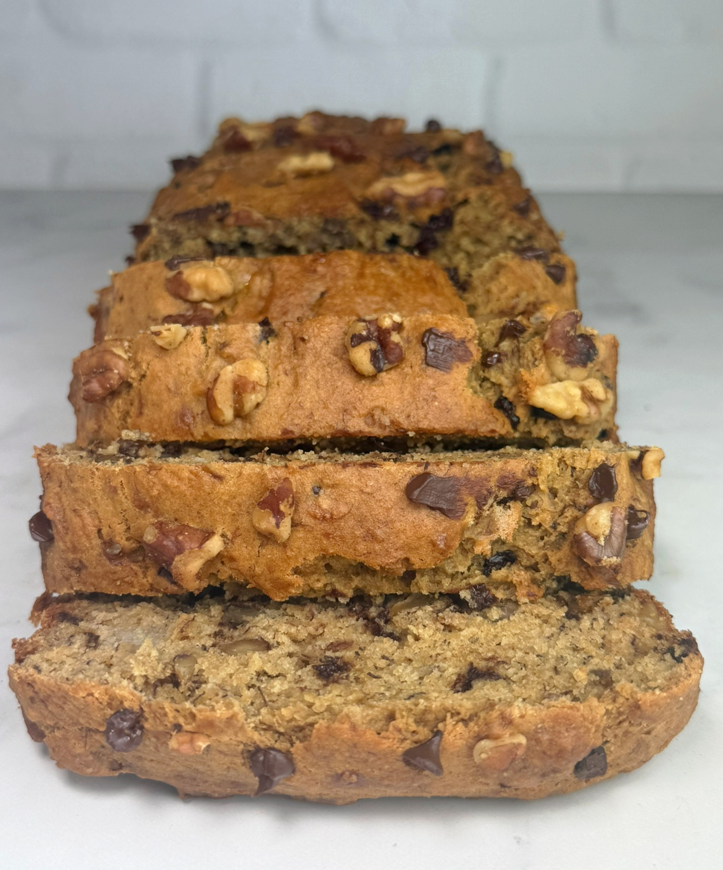 Close-up of stacked slices of whole wheat chocolate chip banana nut bread. The golden-brown crust is topped with walnuts and chocolate chips, and the moist interior shows evenly distributed nuts and melted chocolate throughout.