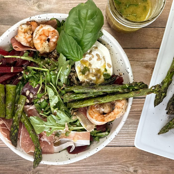 Overhead view of a grilled asparagus, shrimp, burrata, quinoa, arugula, beet, and prosciutto salad topped with sunflower seeds and fresh basil, served with sherry-basil vinaigrette on a wooden table.