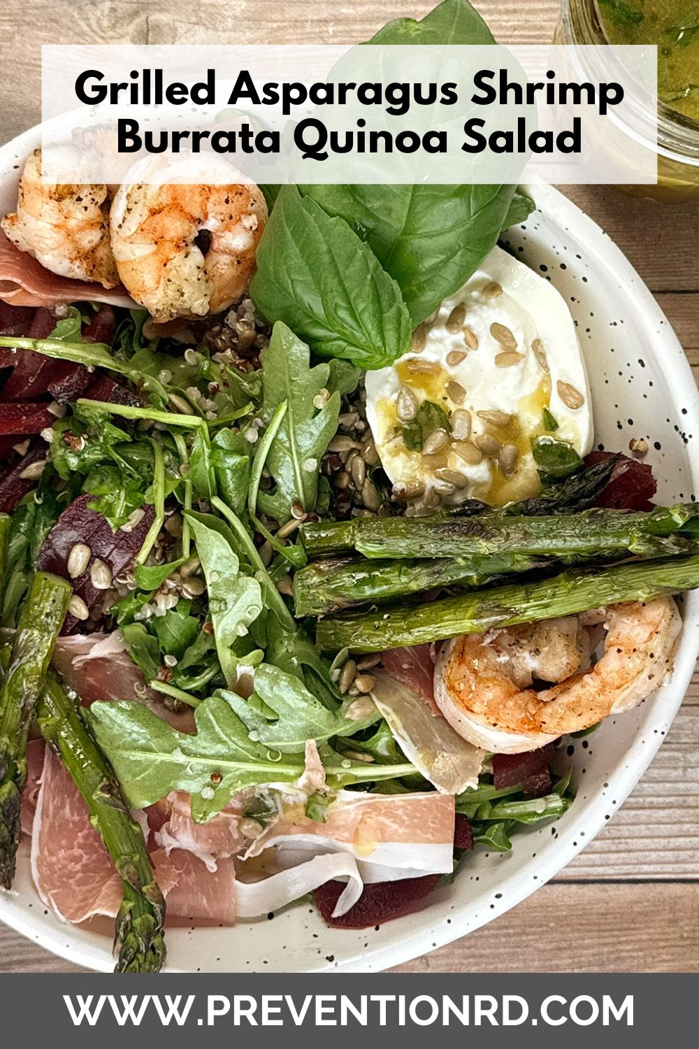 Overhead view of a grilled asparagus, shrimp, burrata, quinoa, arugula, beet, and prosciutto salad topped with sunflower seeds and fresh basil, served with sherry-basil vinaigrette on a wooden table. via @preventionrd