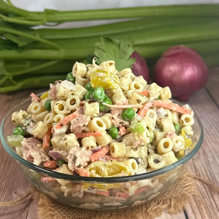 Creamy pesto pasta tuna salad made with small pasta, tuna, peas, shredded carrots, celery, and red onion in a glass bowl with fresh vegetables in the background.