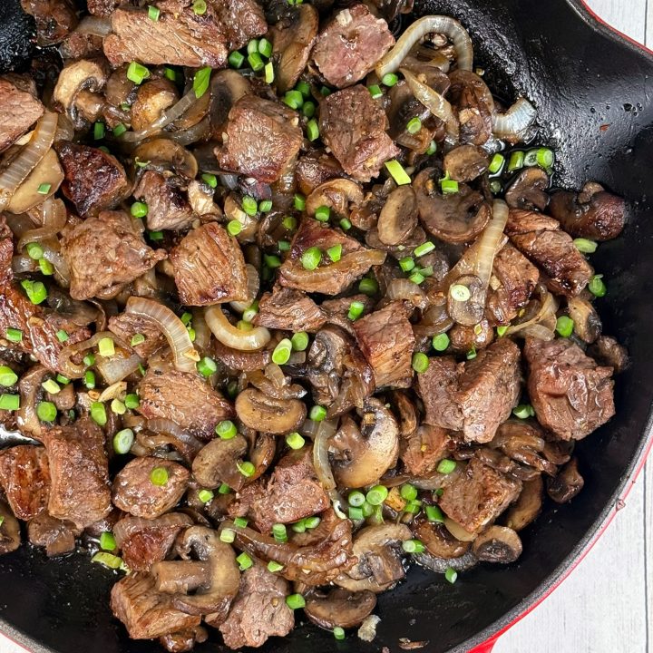 Overhead view of Steak Bites with Horseradish Dipping Sauce featuring seared strip steak, sautéed mushrooms, caramelized onions, and fresh chives in a cast-iron skillet.