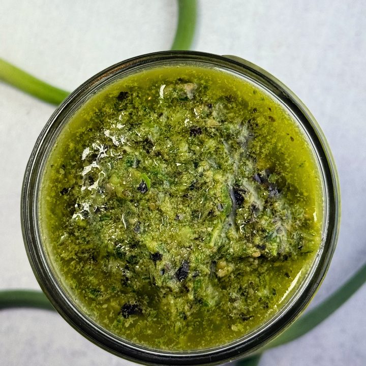 Homemade garlic scape pesto made with fresh garlic scapes from a local CSA, basil, sunflower seeds, lemon juice, and olive oil in a glass jar, photographed from above.